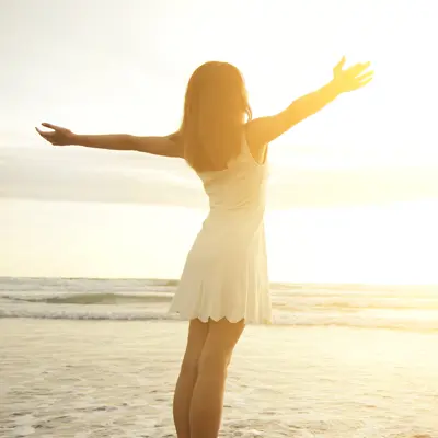 Woman standing on a sunny beach with arms raised and open toward the ocean